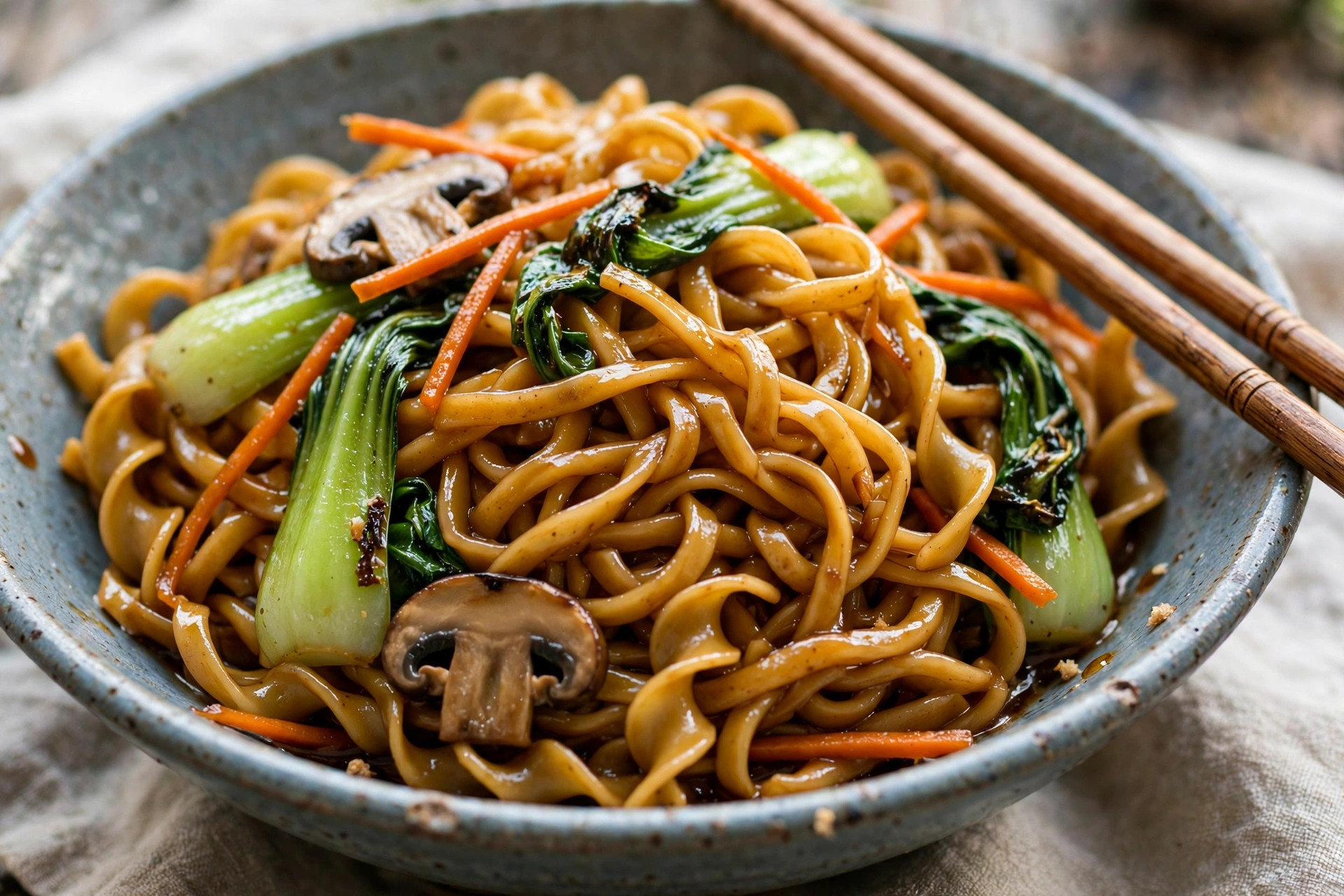 A bowl of lo mein with soft glossy thick noodles, bok choy, mushrooms, carrots and chopsticks
