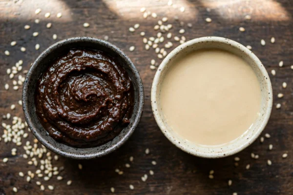 Two ceramic bowls side by side holding dark Chinese sesame paste and pale tahini with scattered sesame seeds